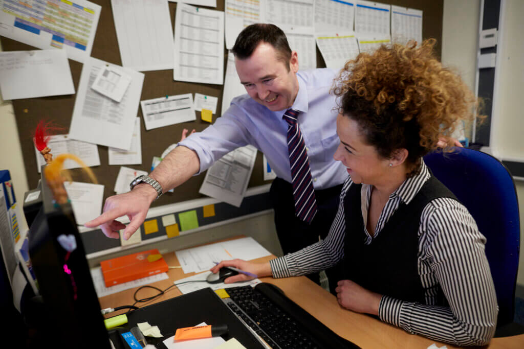 At a desk in the school office, a man points at the computer, whilst a woman looks at the screen.