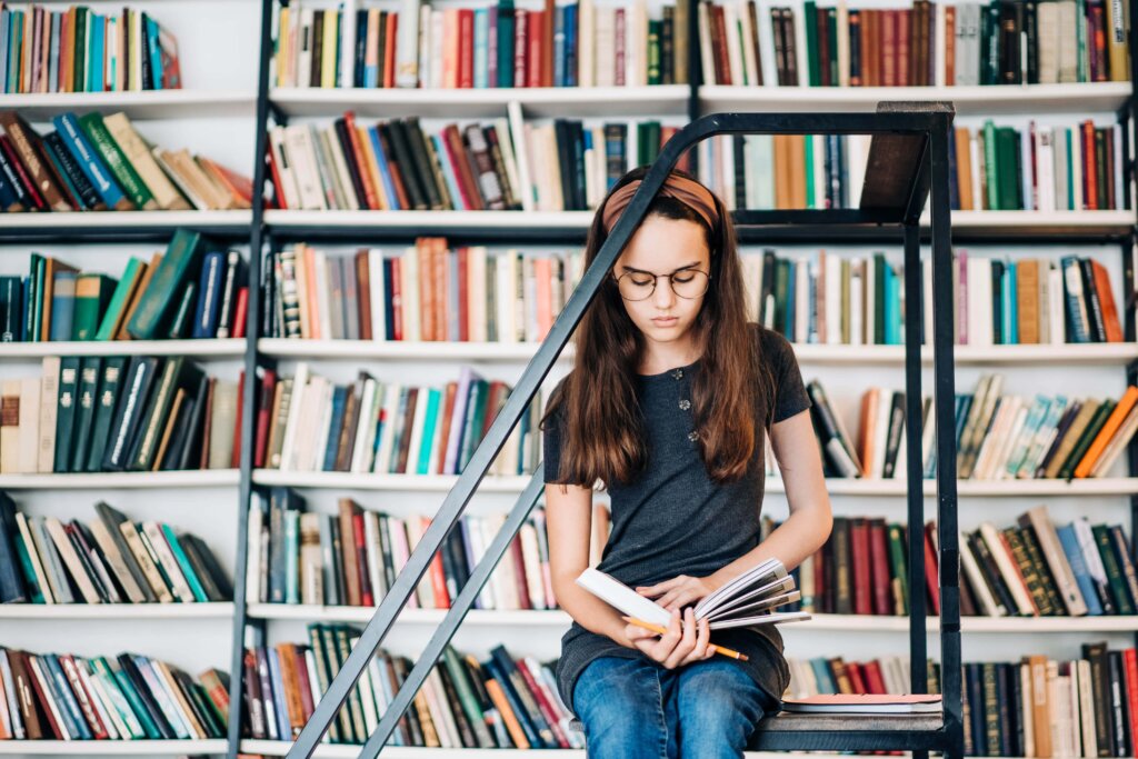 A girl reading a book, sitting in front of shelves of books.