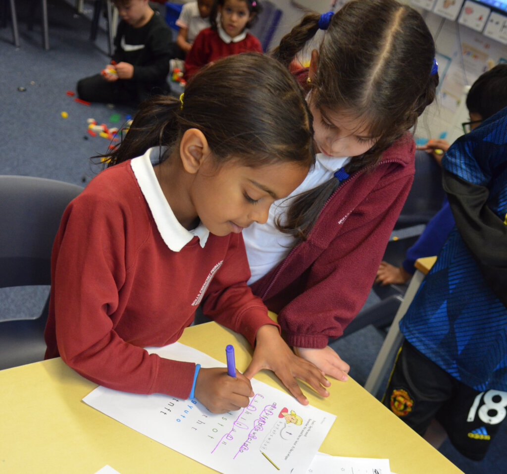 A young school girl writing with a coloured pen, her friend standing beside her.