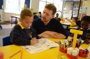 A teacher speaking to a young pupil as they write in their class book.