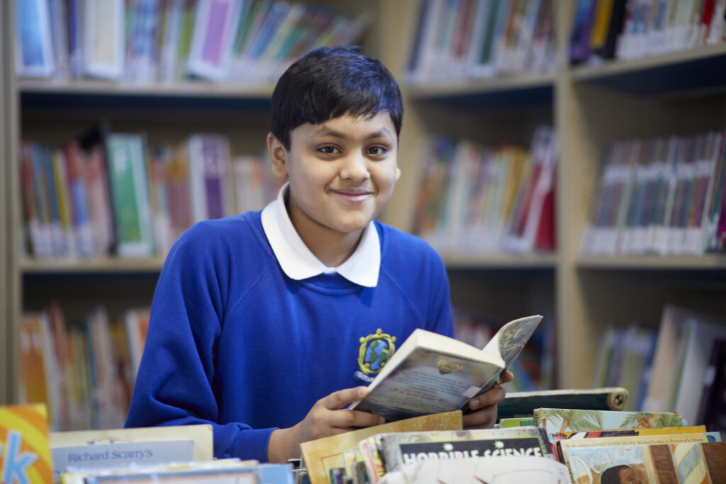A boy smiling in the library, holding an open book in his hands.