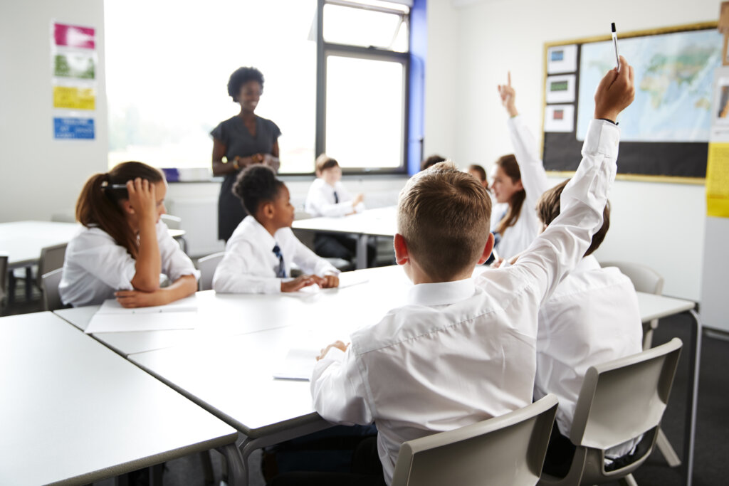 High School Students Wearing Uniform Raising Hands To Answer Question Set By Teacher In Classroom