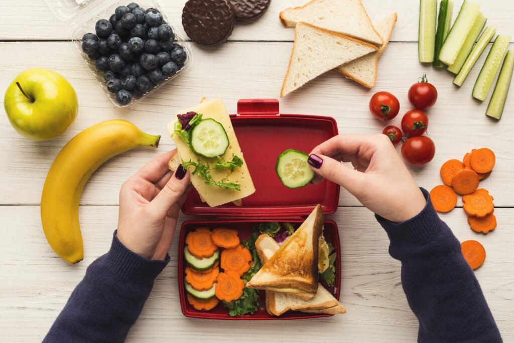 A pair of hands taking cucumber out of an open lunchbox, where there are sliced vegetables and bread. Around the lunchbox, there are grapes, bread and a banana on the table.
