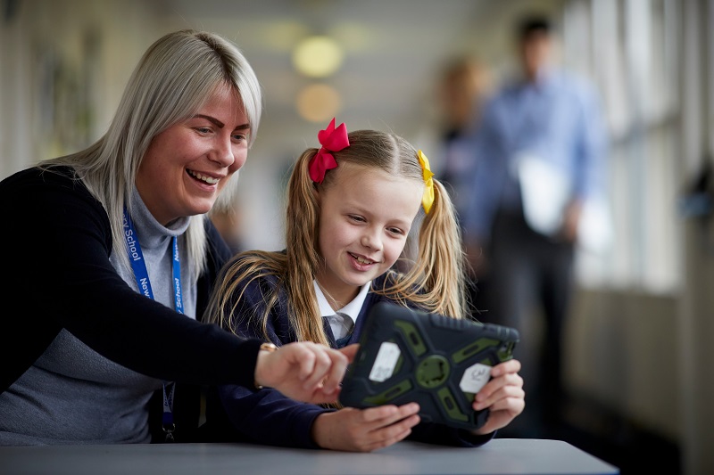 A girl holding a tablet, whilst a teacher sitting beside her points towards the screen.