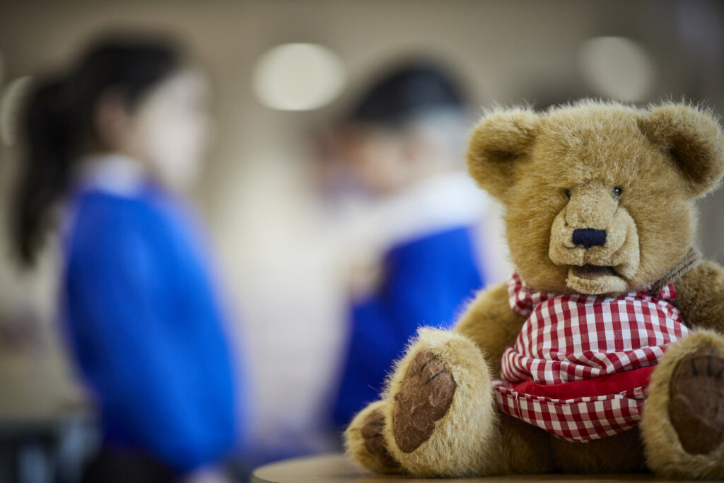 A teddy bear, with two pupils in the background out of focus.