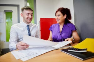 A man and woman in a business setting, sitting at a desk and looking at documents.