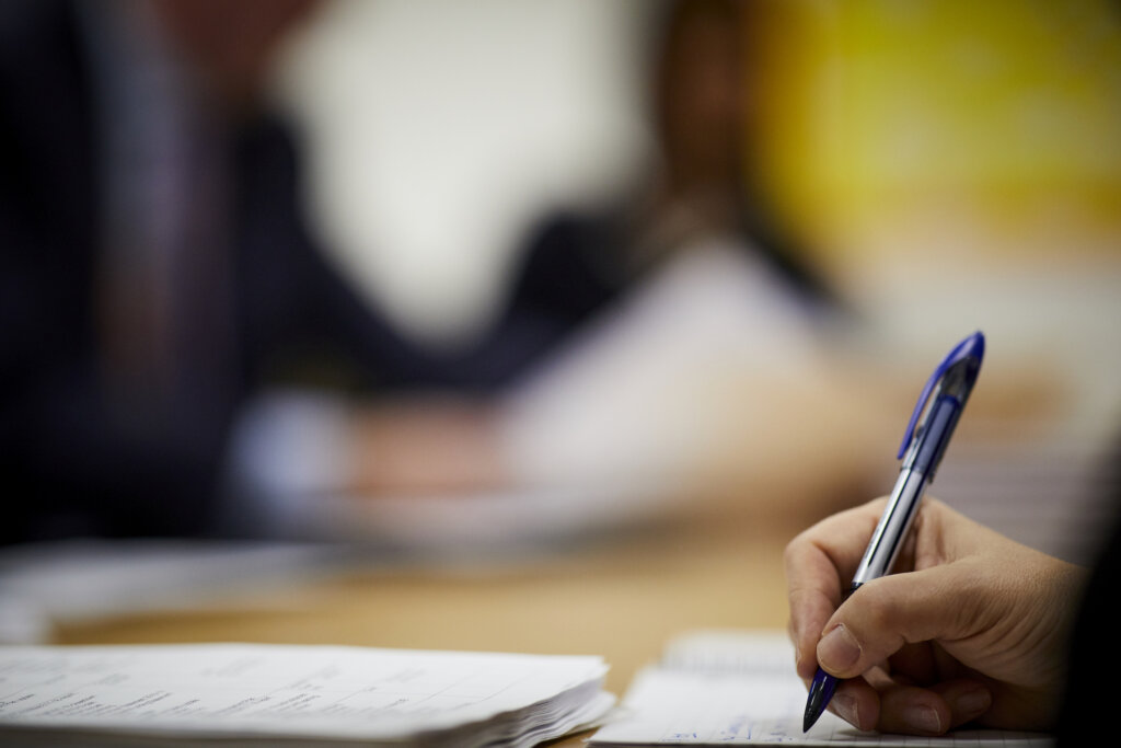 A woman's hand writing with a pen, a stack of paper on the table next to her.