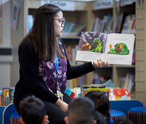 A teacher reads a picture book, as young children sit on the floor and listen.