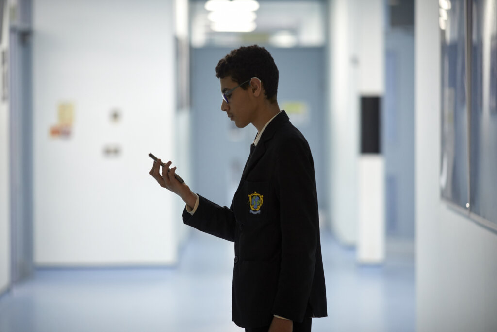 A teenage boy standing alone in a school corridor, looking at his phone.