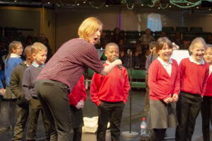 School children stand together on stage, a music teacher holds a microphone towards a boy.