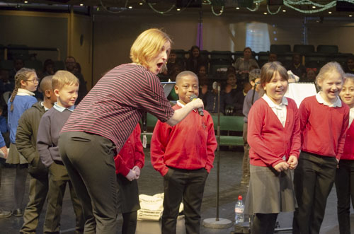 School children stand together on stage, a music teacher holds a microphone towards a boy.