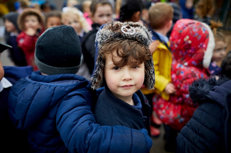 A young boy wearing a hat, standing in the playground with his friends, and looking at the camera behind him.