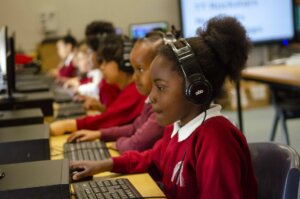 A girl sits in front of a computer with headphones on, other school children sit at their computer stations behind her.