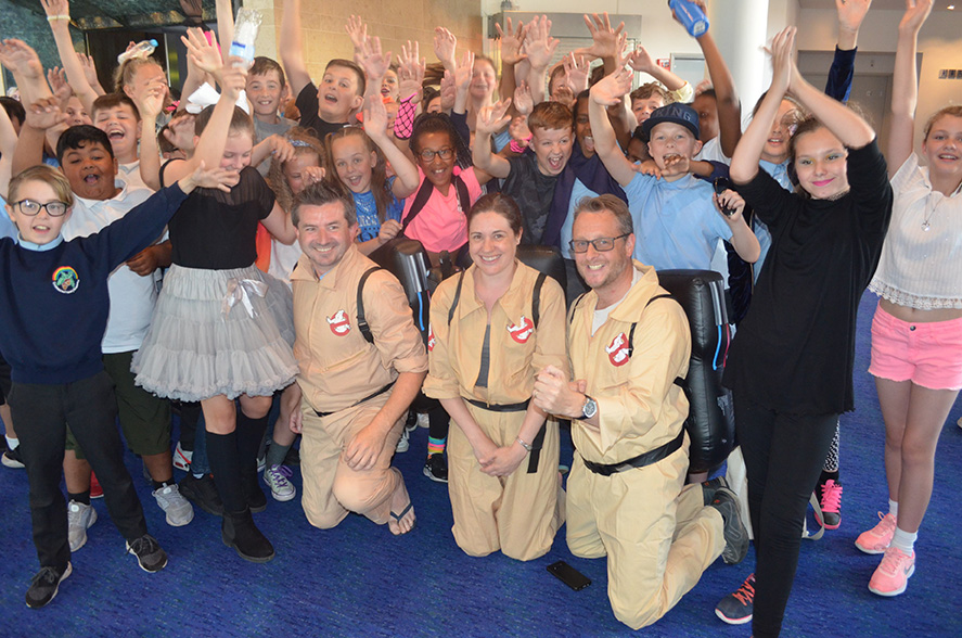 School children cheering, dressed up in 80s costumes with school staff dressed as ghostbusters.