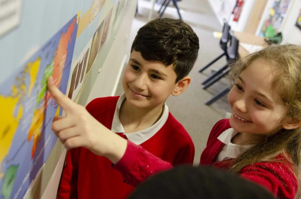 Two children looking at a classroom display, the girl points to a world map as the boy looks on