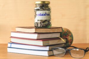 A jar of coins, labelled "education," sitting on a stack of books, beside a pair of glasses and a globe.