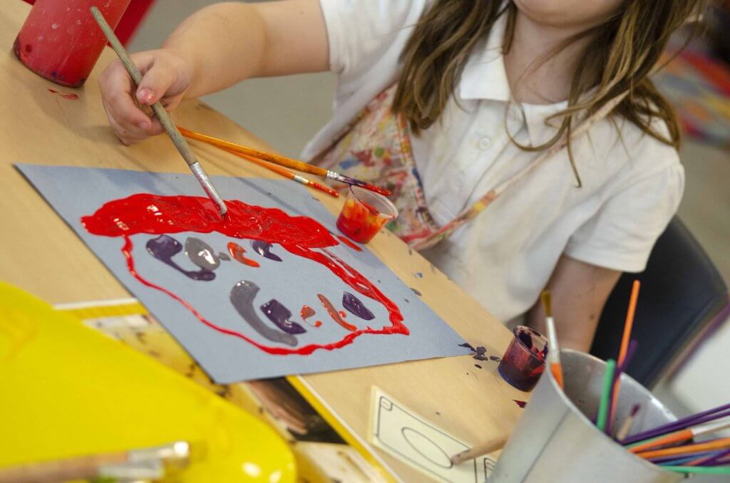 A young girl painting a picture with a paint brush and red paint.