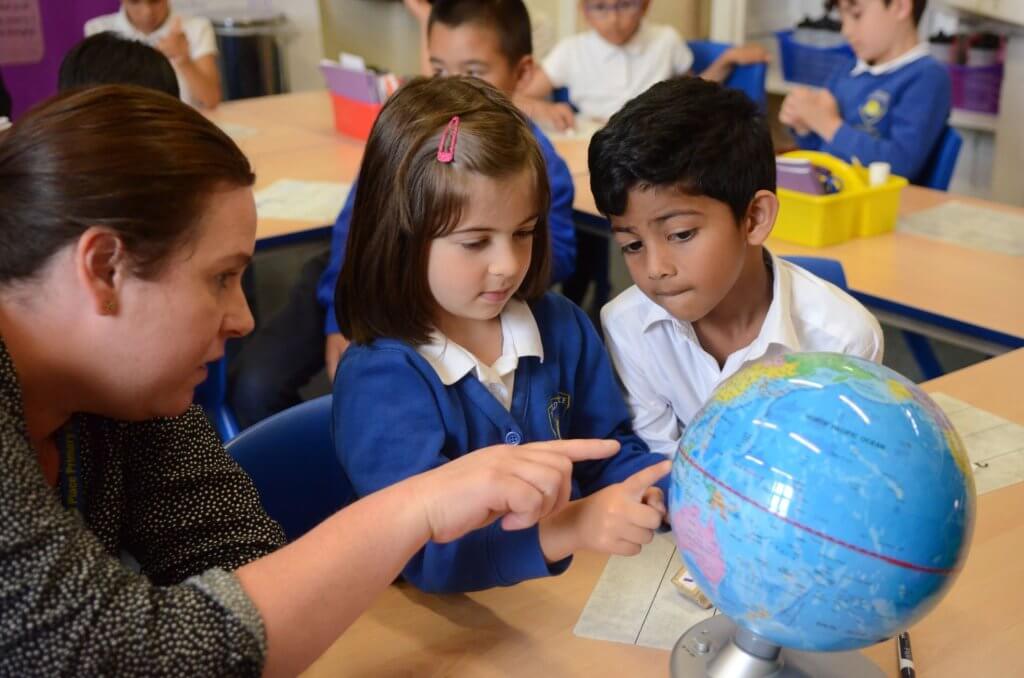 A teacher, with a young girl and boy, looking at a globe on a desk in the classroom.