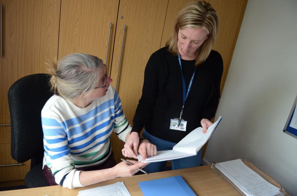 In an office, two women looking at the pages of a book.