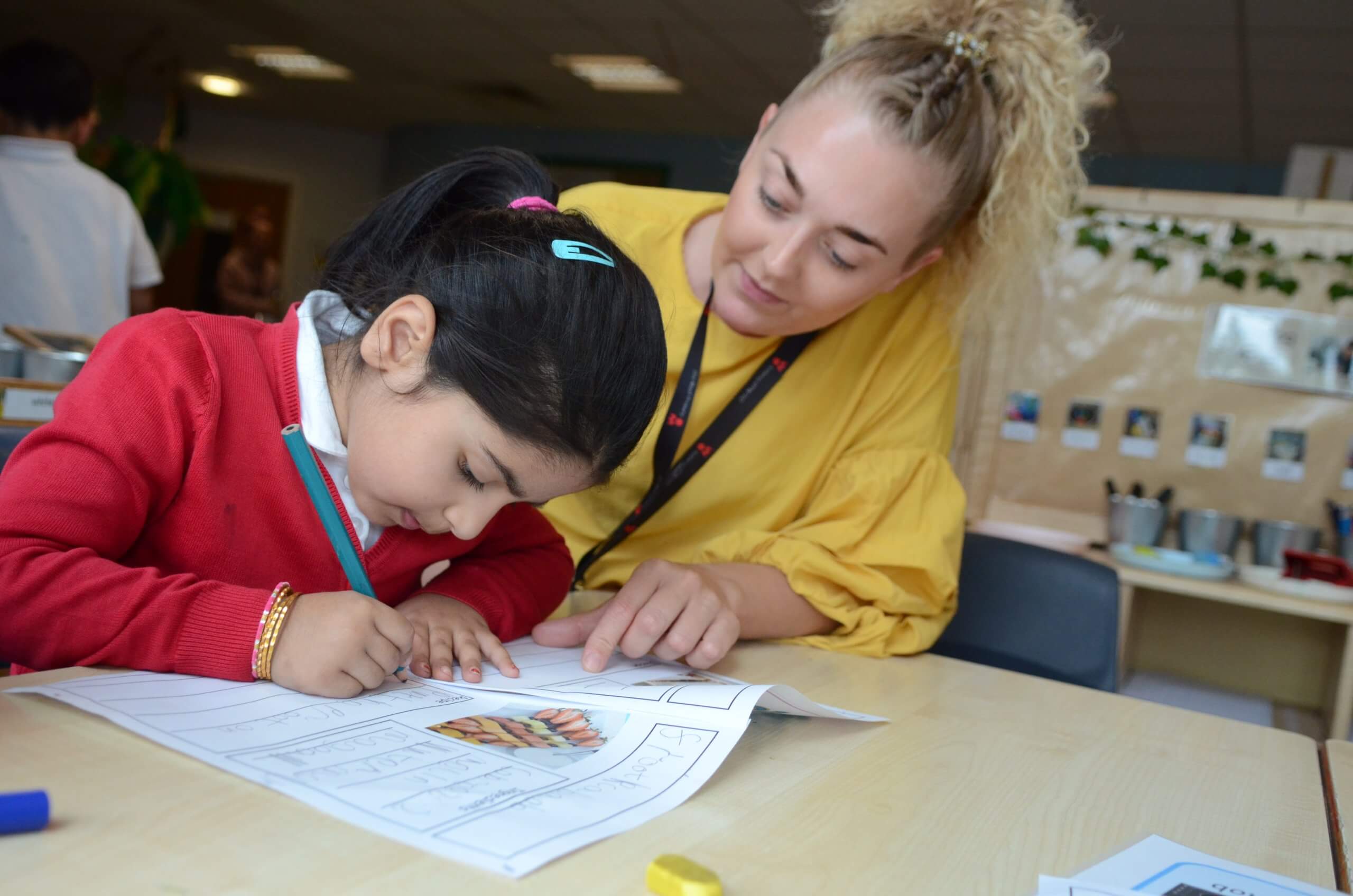 A young girl writing on a piece of paper and a teacher watching over her