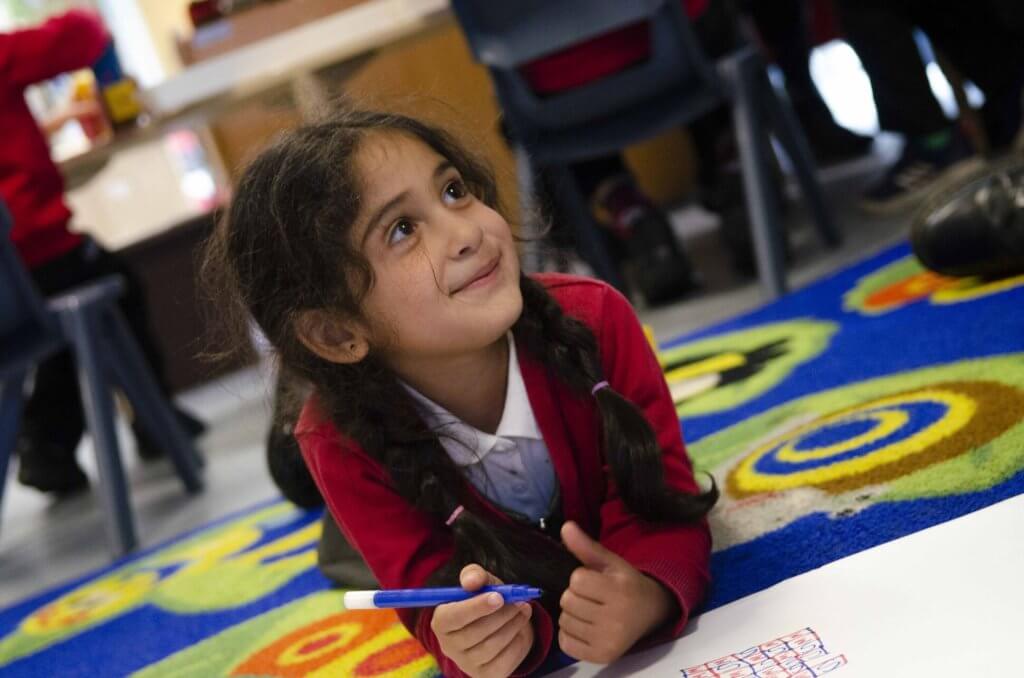 A girl in the early years classroom, lying on the play mat with a pencil and paper.