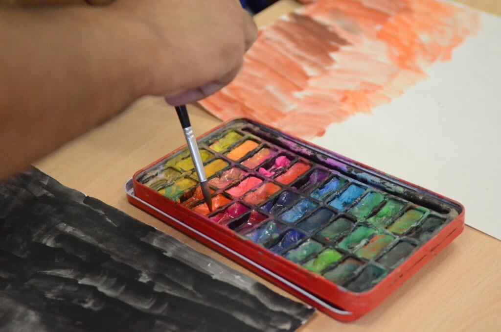 Close up of hand dipping paintbrush into box of coloured paints.