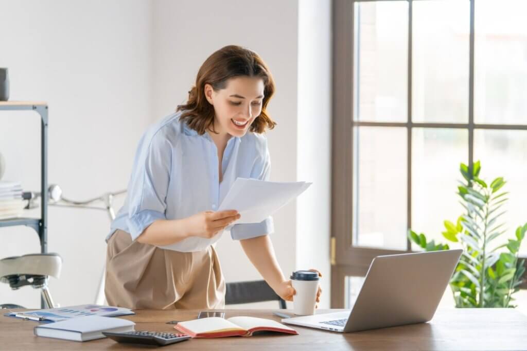 A woman standing at a desk, holding a piece of paper, looking down at her laptop.