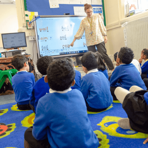 Teacher at the front of the early years classroom, pointing to the whiteboard, whilst children sit on the floor looking on.