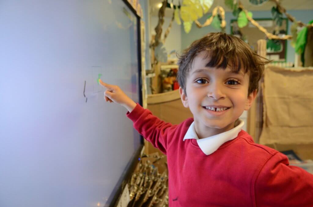 A young school boy smiling as he points to the whiteboard.